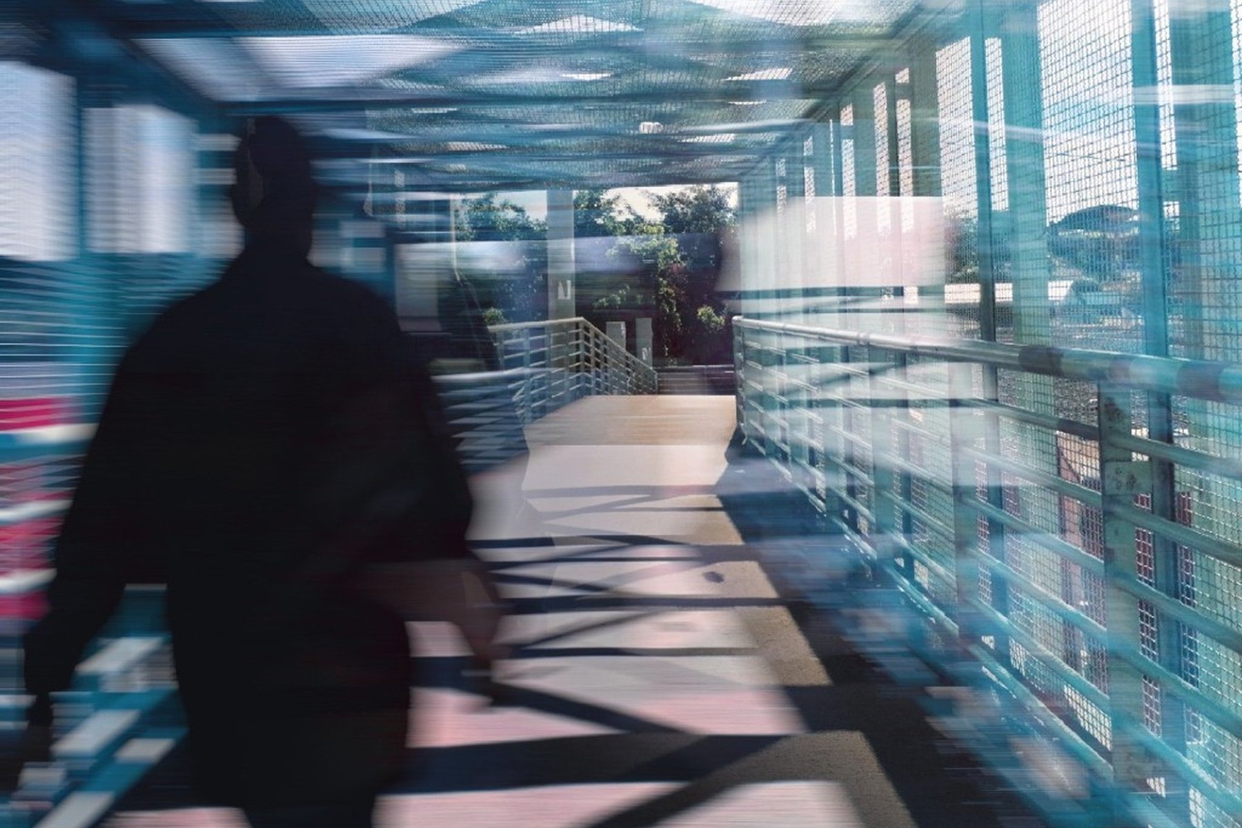 A figure walking a teal mesh pedestrian bridge, streaked by motion blur